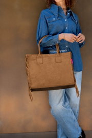 Braune Wildleder-Optik Shopper-Tasche mit langen Henkeln und Fransen an den Seiten, getragen von einer Frau im Jeanshemd und hellen Jeans vor braunem Hintergrund, Frontansicht in leichter Seitenperspektive.