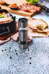 Doppelmühle DUOMILL MINI aus Edelstahl, gefüllt mit Salz und Pfeffer, auf einem Tisch mit Sandwiches und Gewürzen, aus der Vogelperspektive fotografiert.