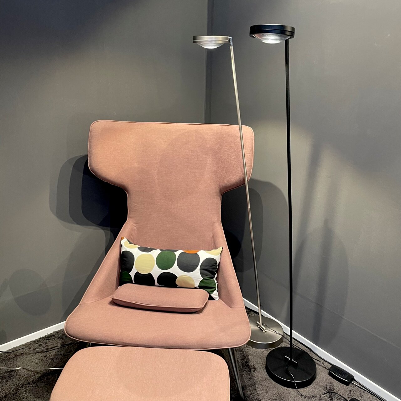 Zwei moderne LED-Stehleuchten, eine silberne und eine schwarze, stehen neben einem rosa Sessel mit einem bunten Kissen. Perspektive: seitlich.
