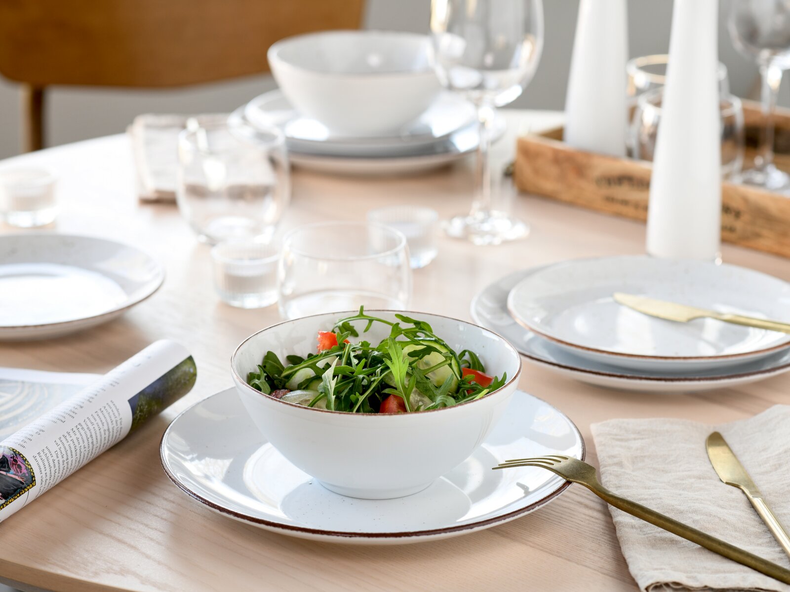 Weißes Keramik-Schüssel- und Teller-Set mit braunem Rand, vorn im Fokus mit frischem Rucola-Salat auf gedecktem Esstisch, perspektivisch in leichter Schrägansicht von vorne oben.