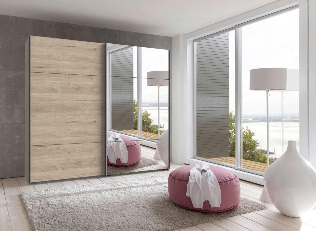 Z2 Schwebetürenschrank 2-türig ERNIE Moderner Schwebetürenschrank mit zwei Türen, eine in Holzoptik und die andere verspiegelt, in einem hellen Raum mit großen Fenstern und Blick auf das Wasser. Perspektive von vorne.
