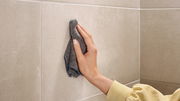 Eine Hand reinigt eine beige geflieste Wand mit einem grauen Tuch aus der Perspektive von vorne.