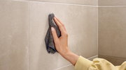 Eine Hand reinigt eine beige geflieste Wand mit einem grauen Tuch aus der Perspektive von vorne.