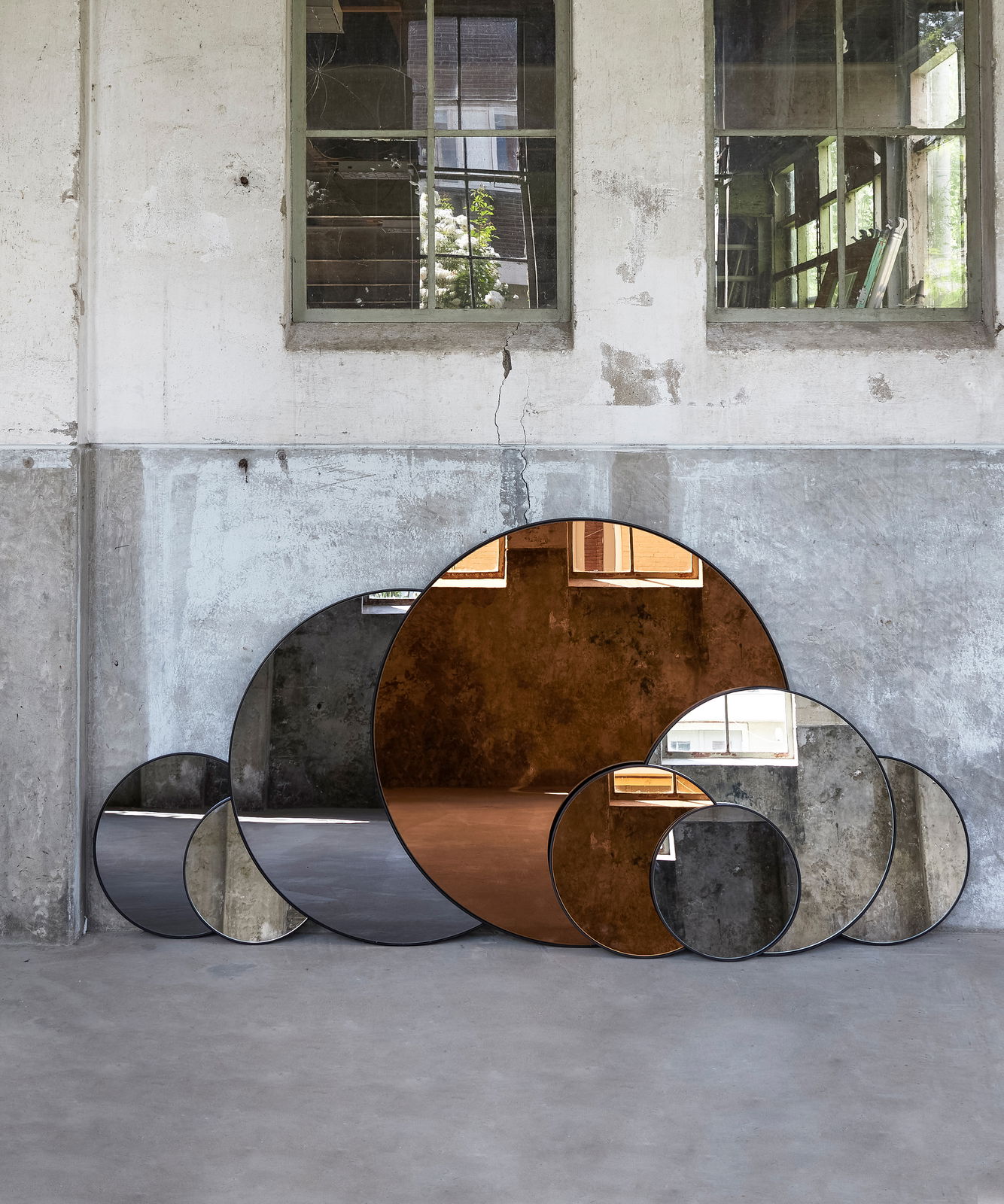 Light&Living Spiegel ESPEJO Mehrere runde Spiegel in verschiedenen Größen, die an einer Wand in einem industriellen Raum lehnen. Die Spiegel reflektieren die Umgebung und erzeugen ein modernes, künstlerisches Ambiente. Perspektive: Frontalansicht.