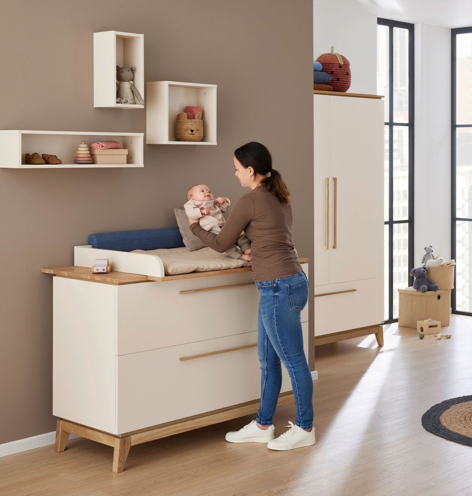 Frau steht vor einer Wickelkommode und hält ein Baby, im Hintergrund sind Wandboxen an der Wand montiert, seitliche Perspektive