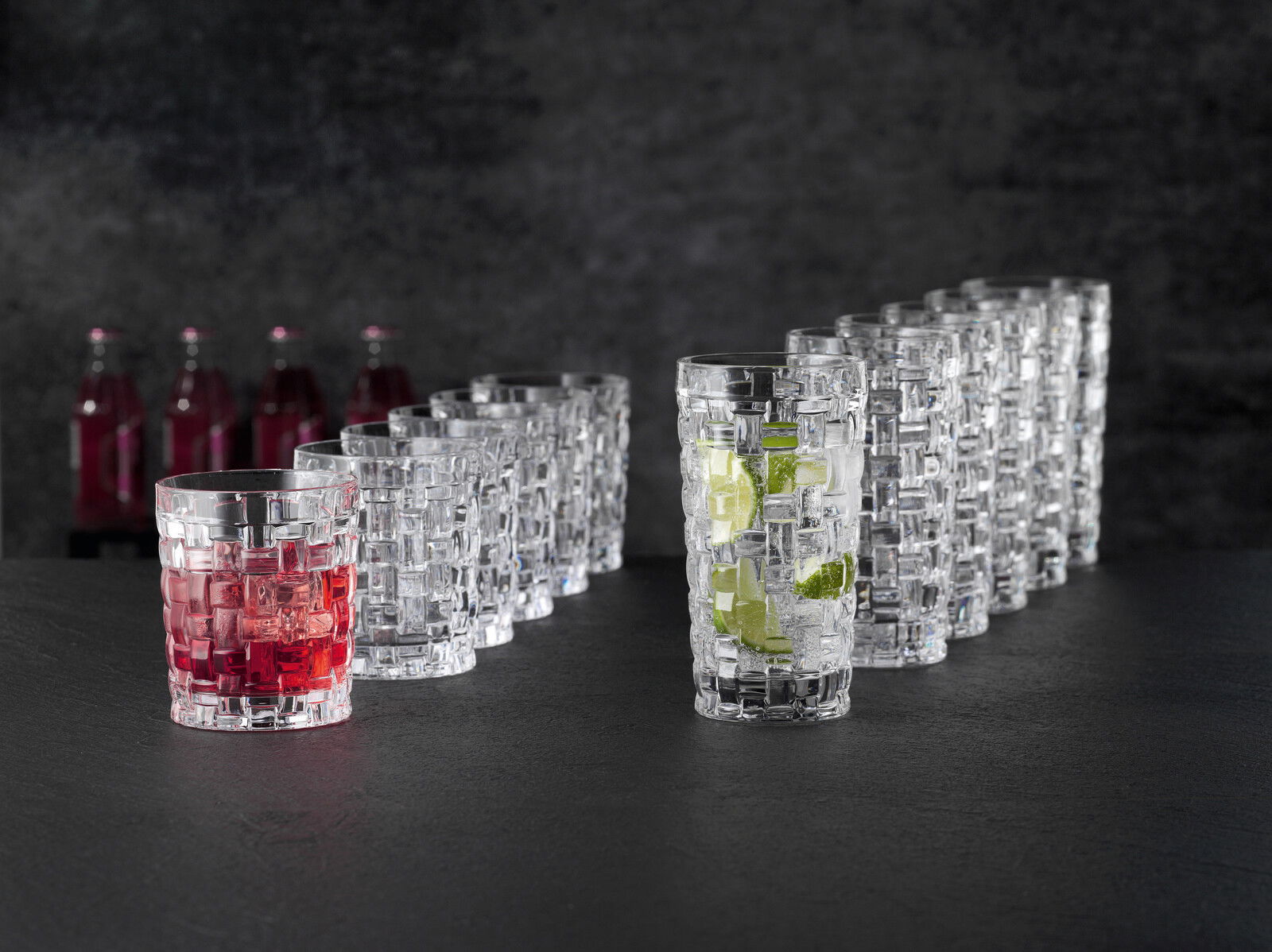 Ein Set von zwölf Bossa Nova Bechern, stilvoll auf einem dunklen Hintergrund arrangiert. Die Becher haben ein elegantes, geflochtenes Design. Zwei der Becher sind mit Getränken gefüllt, einer mit rotem und der andere mit klarem Getränk und Limettenstücken
