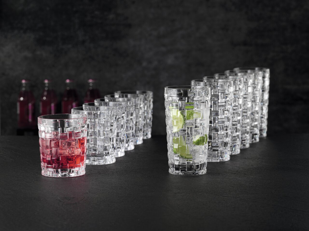 Ein Set von zwölf Bossa Nova Bechern, stilvoll auf einem dunklen Hintergrund arrangiert. Die Becher haben ein elegantes, geflochtenes Design. Zwei der Becher sind mit Getränken gefüllt, einer mit rotem und der andere mit klarem Getränk und Limettenstücken