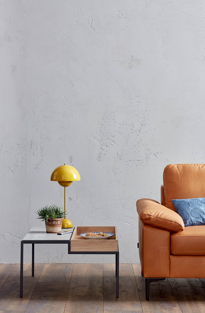 Teilansicht eines modernen Sofas in Lederoptik mit einem blauen Kissen, daneben ein Beistelltisch mit gelber Lampe und Pflanze, aus seitlicher Perspektive.