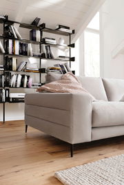 Beige Longchairkombination in einem modernen Wohnzimmer, seitliche Perspektive mit Bücherregal im Hintergrund.