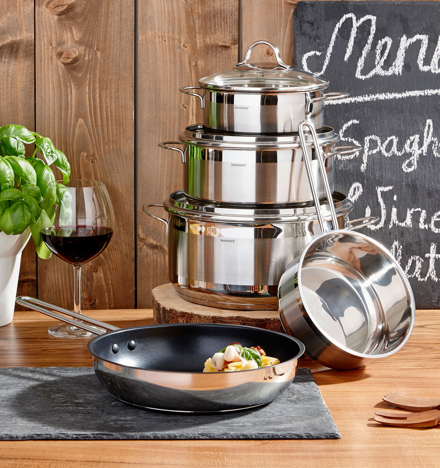 Perspektive: Frontalansicht auf ein 5-teiliges Topfset aus Edelstahl, bestehend aus gestapelten Töpfen mit Deckeln und einer Pfanne im Vordergrund, dekoriert mit frischen Kräutern und einem Glas Rotwein.