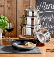 Perspektive: Frontalansicht auf ein 5-teiliges Topfset aus Edelstahl, bestehend aus gestapelten Töpfen mit Deckeln und einer Pfanne im Vordergrund, dekoriert mit frischen Kräutern und einem Glas Rotwein.