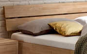 Holz-Kopfteil eines Bettes mit zwei Kissen, eines in Grau und eines in Gold, aus seitlicher Perspektive