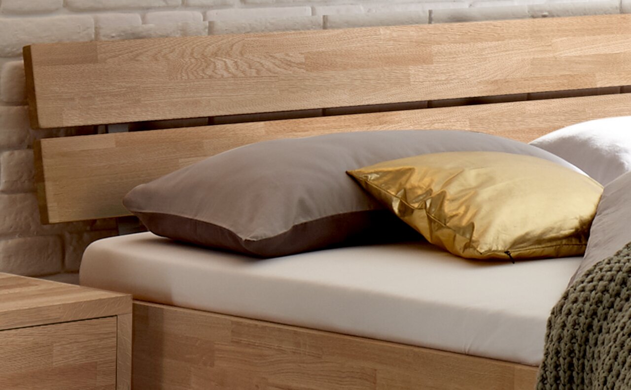 Holz-Kopfteil eines Bettes mit zwei Kissen, eines in Grau und eines in Gold, aus seitlicher Perspektive