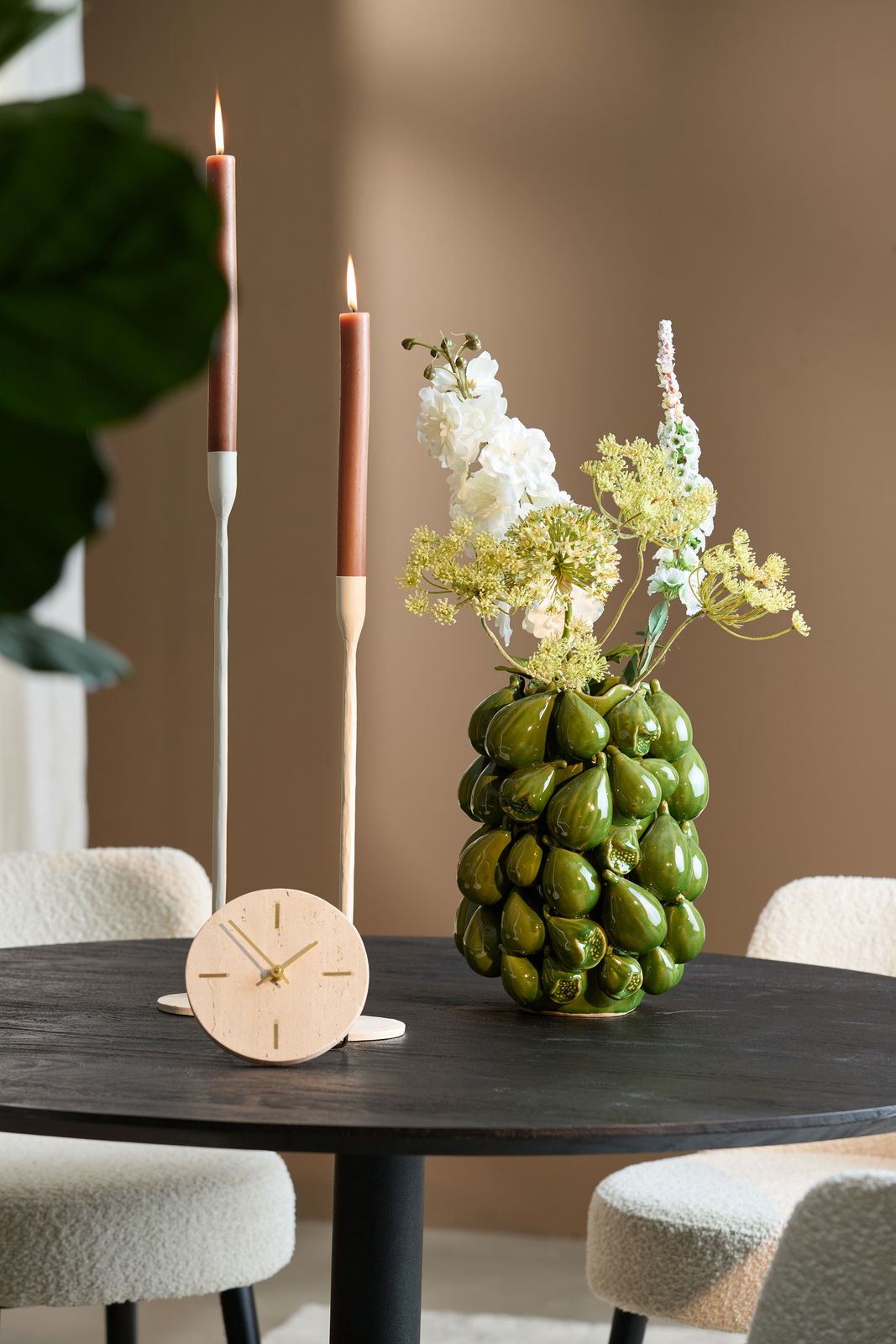 Runde Tischuhr aus hellem Holz mit goldenen Zeigern, auf einem dunklen Holztisch platziert. Im Hintergrund sind zwei brennende Kerzen und eine grüne Vase mit Blumen zu sehen. Perspektive: seitlich von vorne.