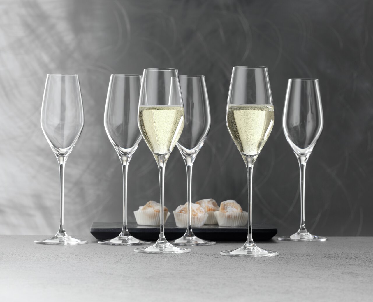 Sechs elegante Champagnerkelche der Serie Topline, zwei gefüllt mit Champagner, auf einer grauen Oberfläche mit einem modernen Hintergrund. Perspektive von vorne.