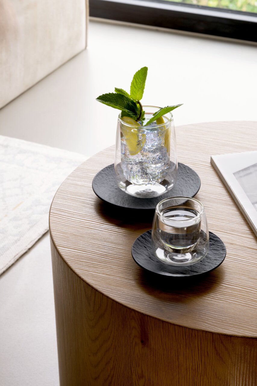 Twee espressoschoteltjes uit de Manufacture-serie op een houten tafel, met glazen gevuld met water en munt, gefotografeerd vanuit een zijaanzicht.