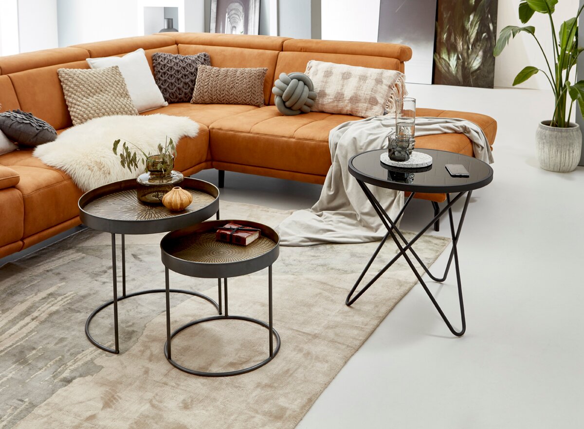 Wohnzimmer-Szene mit cognacfarbenem Ecksofa, Kissen und Decke sowie drei schwarzen Beistelltischen mit Tablett-/Glasplatte auf hellem Teppich, fotografiert aus leicht erhöhter Frontperspektive.