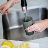 Eine Hand hält einen grauen Becher unter einen Wasserhahn, während Wasser in den Becher fließt. Im Vordergrund sind Zitronenscheiben zu sehen. Perspektive von oben.