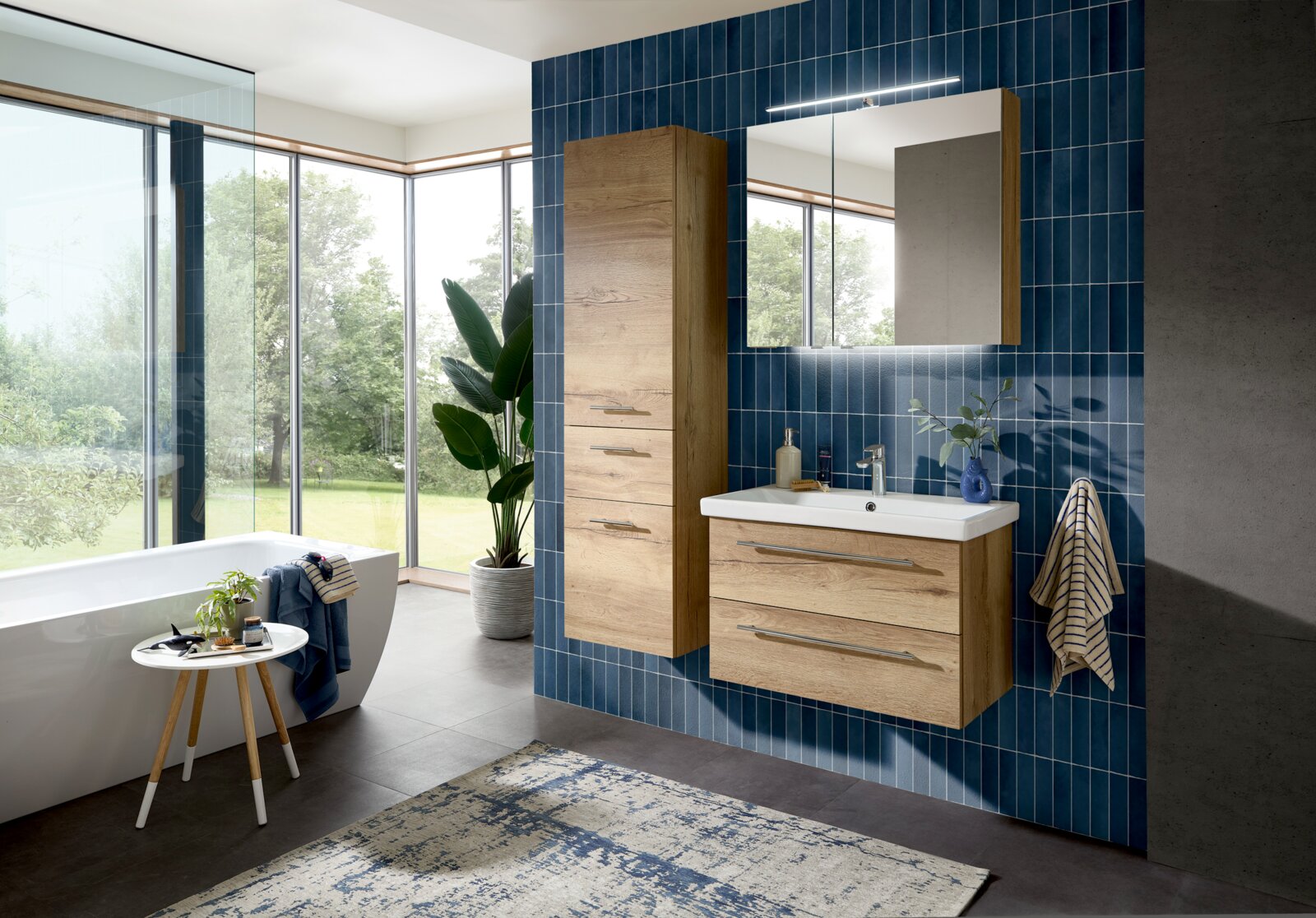 Modernes Badezimmer mit hängendem Waschtisch und Hochschrank in Holzoptik, weißem Waschbecken, Spiegelschrank mit LED-Leuchte vor blauen Fliesen; Perspektive: frontale, leicht seitliche Ansicht auf die Badmöbel.
