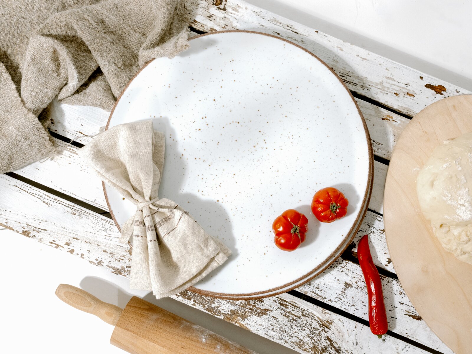 Runder, weiß gesprenkelter Keramik-Essteller mit braunem Rand auf rustikalem, weiß gestrichenem Holztisch, dekoriert mit Stoffserviette und zwei kleinen Tomaten; Perspektive: Draufsicht (Top-Down)