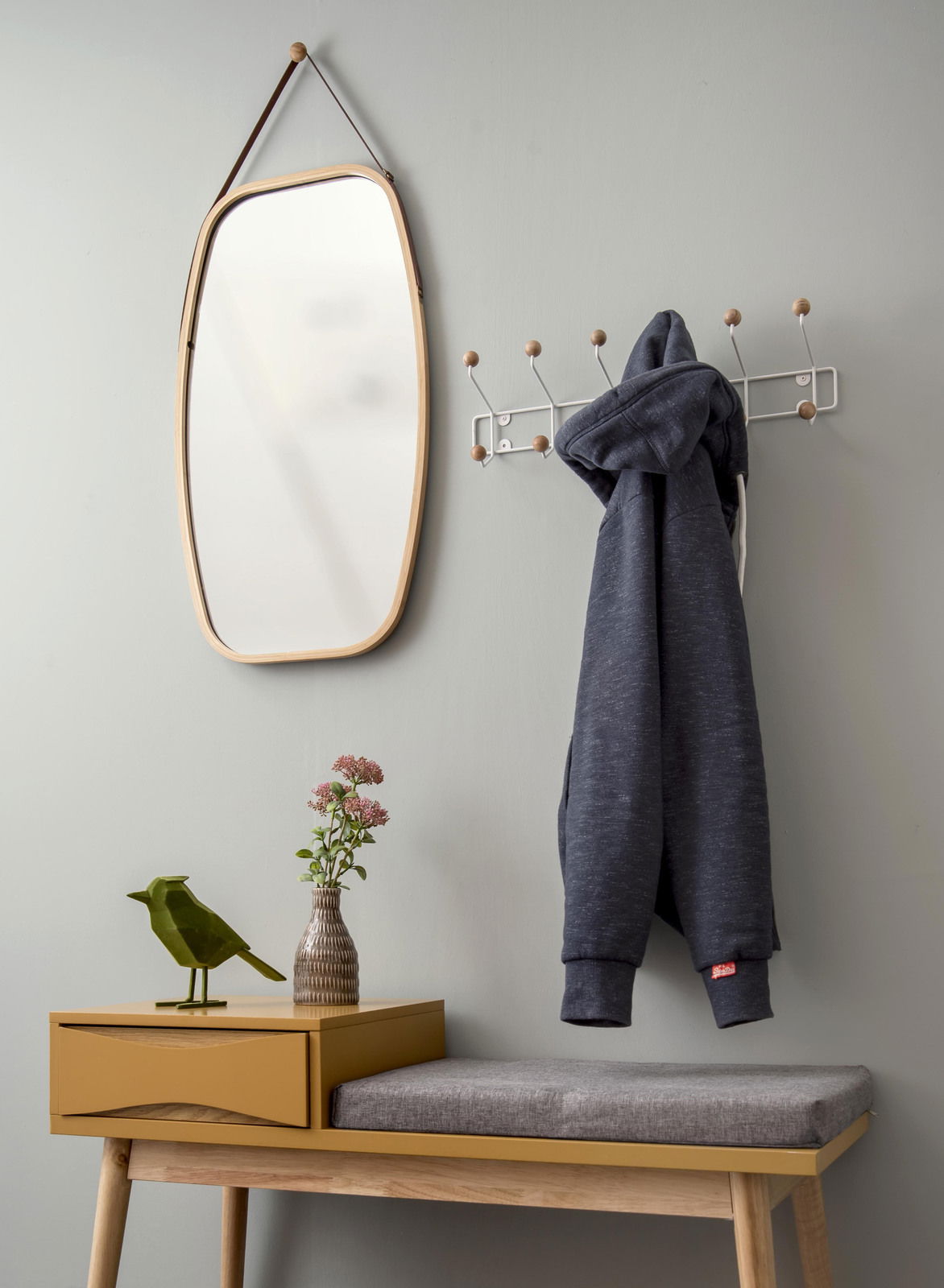 Garderobe mit Spiegel und Hakenleiste, seitliche Perspektive. Ein grauer Hoodie hängt an der Hakenleiste, daneben ein Spiegel mit Holzrahmen. Darunter eine gelbe Bank mit grauem Sitzkissen, dekoriert mit einer Vase und einer Vogelstatue.