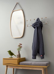 Garderobe mit Spiegel und Hakenleiste, seitliche Perspektive. Ein grauer Hoodie hängt an der Hakenleiste, daneben ein Spiegel mit Holzrahmen. Darunter eine gelbe Bank mit grauem Sitzkissen, dekoriert mit einer Vase und einer Vogelstatue.