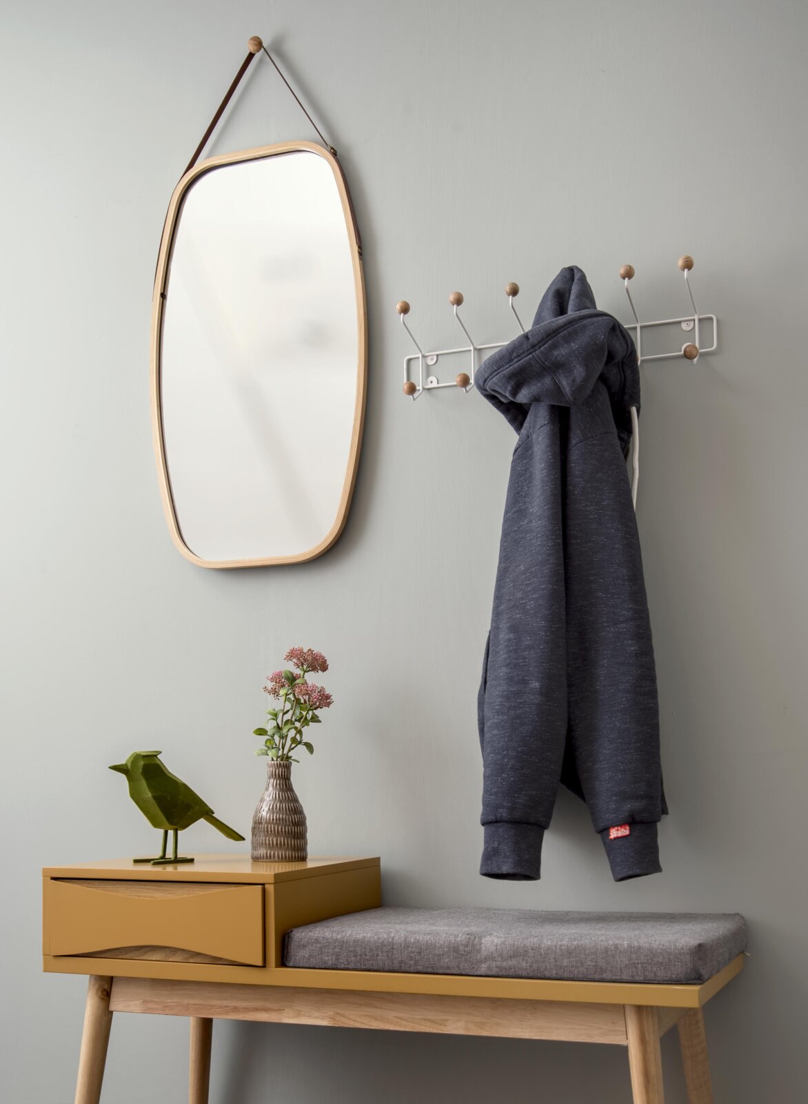 Garderobe met spiegel en kapstok, zijaanzicht. Aan de kapstok hangt een grijze hoodie, ernaast staat een spiegel met houten lijst. Daaronder staat een geel bankje met een grijs zitkussen, versierd met een vaas en een vogelbeeldje.