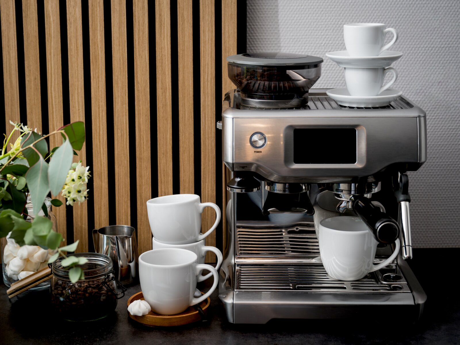 Edelstahl-Espressomaschine mit Bohnenbehälter, weißen Tassen und Zubehör auf Küchenarbeitsplatte, frontal aus leicht schräger Perspektive aufgenommen