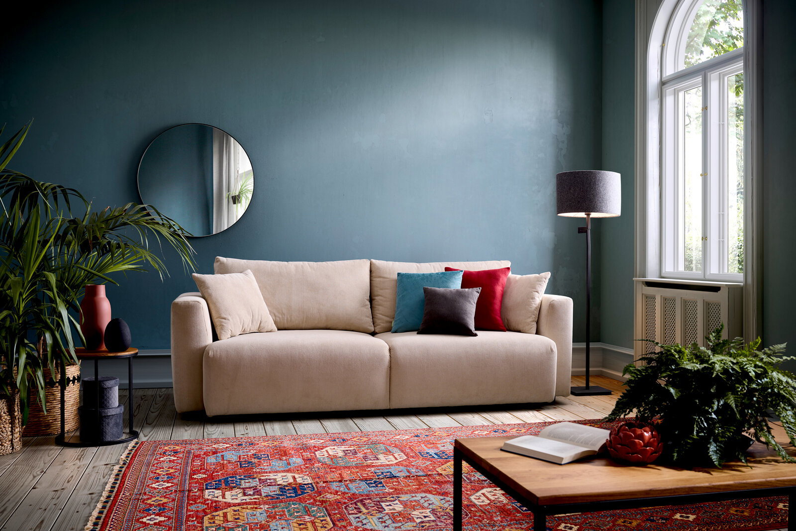 Beiges Funktionssofa PONTE in einem stilvoll eingerichteten Wohnzimmer mit blauen Wänden, dekorativen Kissen und einem runden Spiegel, aus frontaler Perspektive.