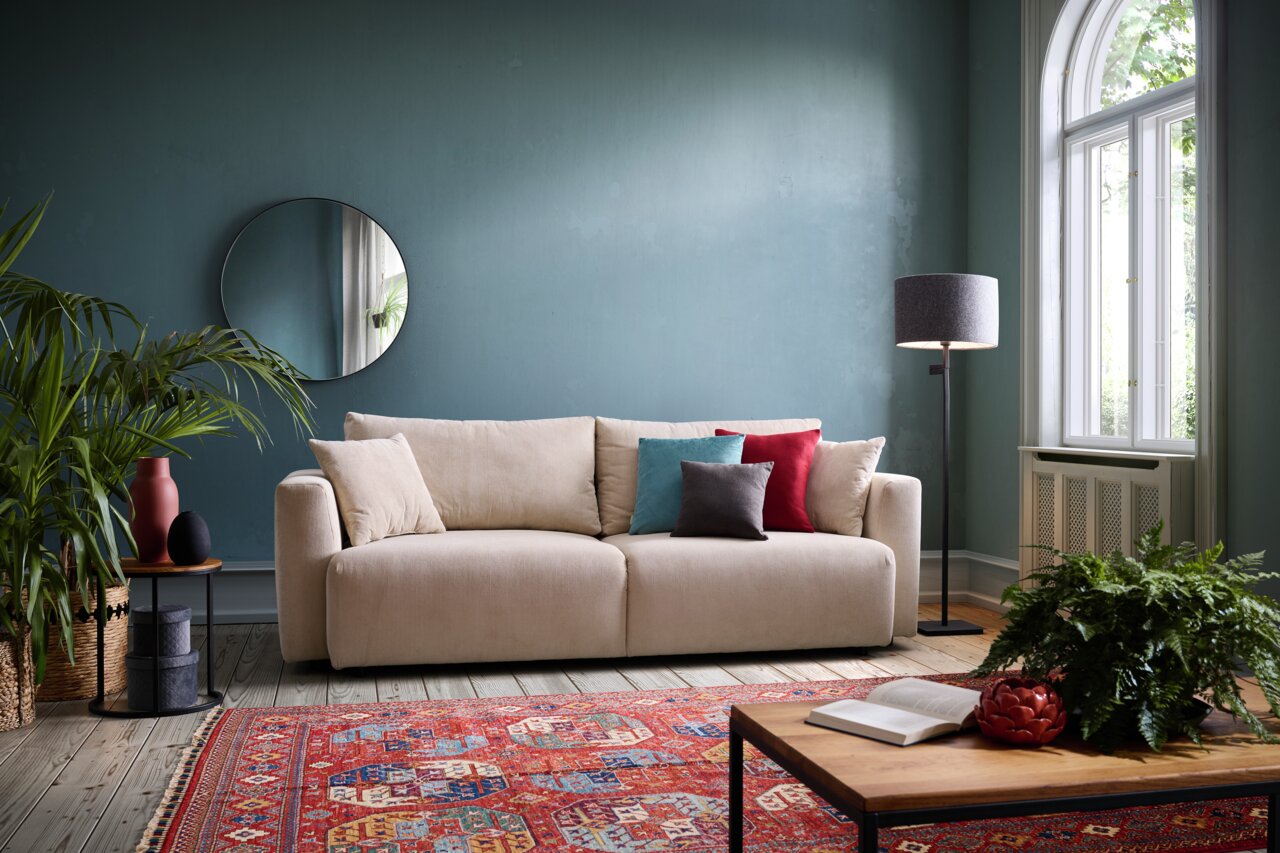 Z2 Schlafsofa PONTE Beiges Funktionssofa PONTE in einem stilvoll eingerichteten Wohnzimmer mit blauen Wänden, dekorativen Kissen und einem runden Spiegel, aus frontaler Perspektive.