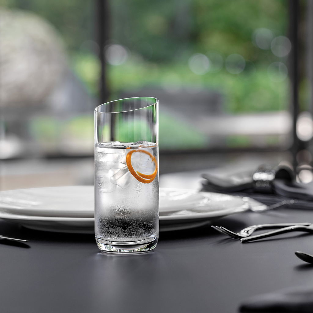 Ein eleganter Longdrinkbecher mit Eiswürfeln und einer Orangenscheibe, auf einem gedeckten Tisch mit unscharfem Hintergrund, aus seitlicher Perspektive.