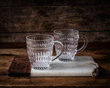 Zwei durchsichtige, strukturierte Becher mit Henkel auf einem rustikalen Holz- und Marmorbrett, seitliche Perspektive.