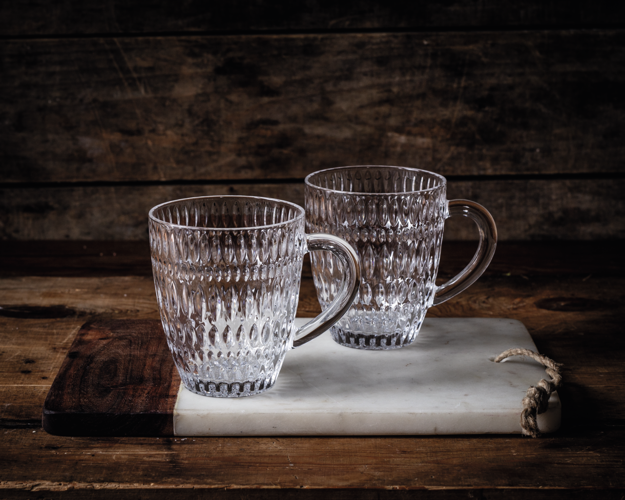 Zwei durchsichtige, strukturierte Becher mit Henkel auf einem rustikalen Holz- und Marmorbrett, seitliche Perspektive.