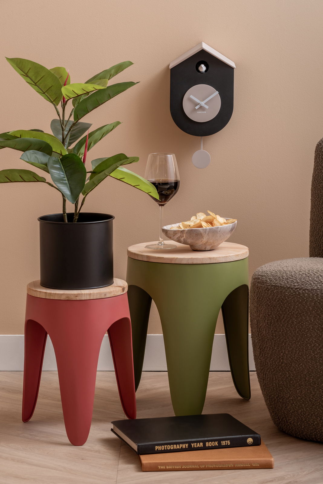 Moderne Wanduhr Grato in Schwarz und Weiß, aufgehängt an einer beigen Wand, mit einem Pendel. Perspektive von vorne, neben dekorativen Elementen wie einem Pflanzenständer und einem Weinglas.