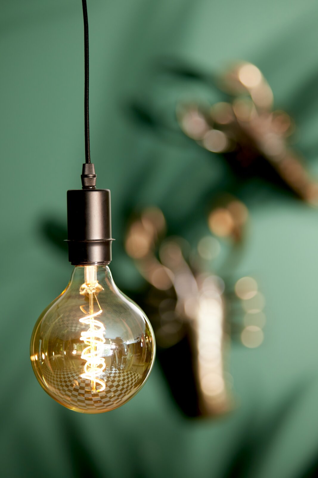 Light&Living Pendelleuchte mit Aufnahme CUP Nahaufnahme einer hängenden Glühbirne mit sichtbarem Glühfaden vor einem unscharfen grünen Hintergrund.