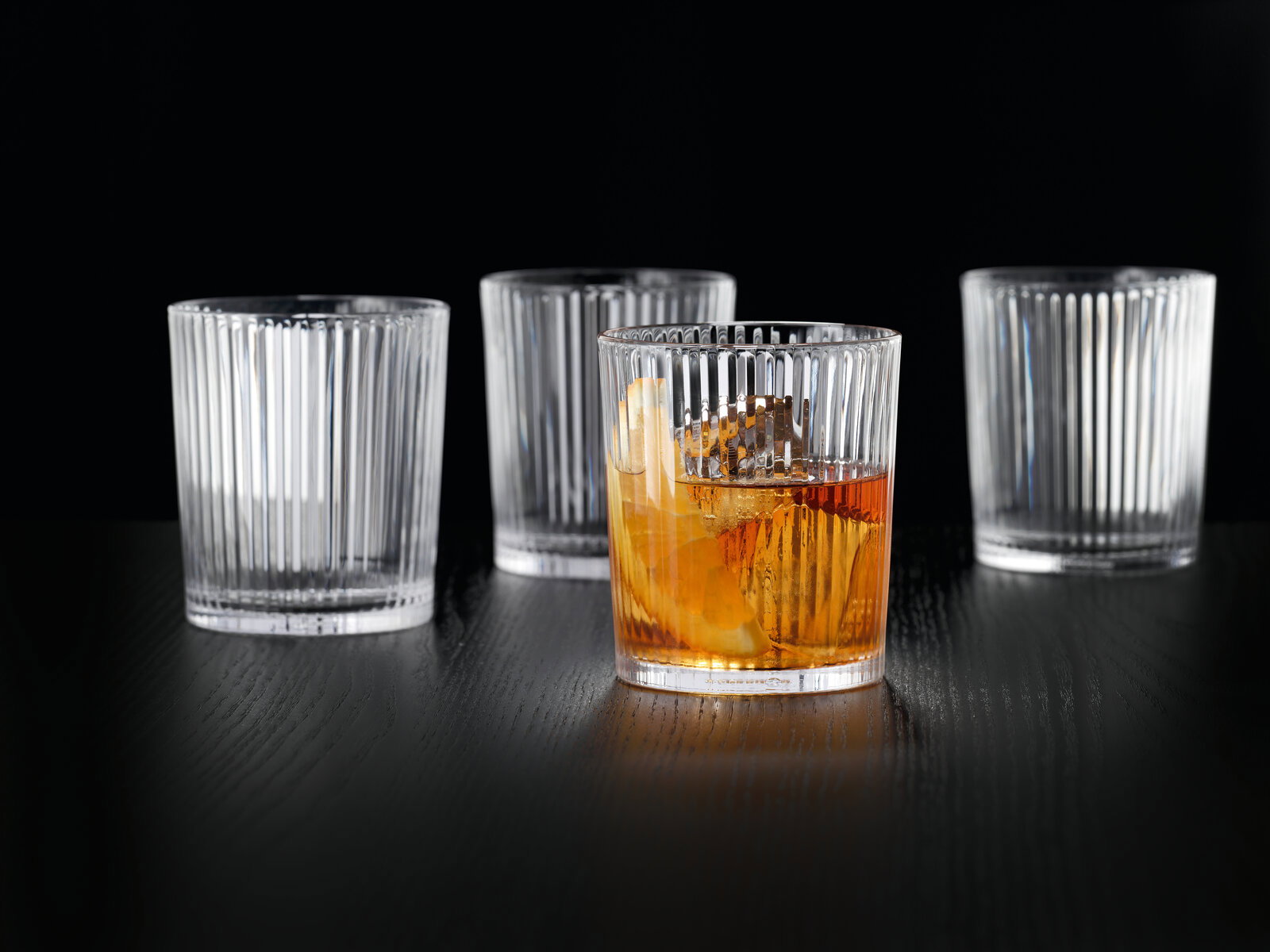 Vier elegante, geriffelte Gläser auf einem dunklen Tisch, eines mit Getränk gefüllt, aus seitlicher Perspektive.