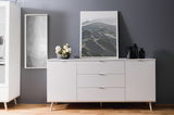 Weißes Sideboard mit drei Schubladen und zwei Türen, dekoriert mit einer Blumenvase und schwarzen Gefäßen, vor einer grauen Wand. Perspektive: Frontalansicht.