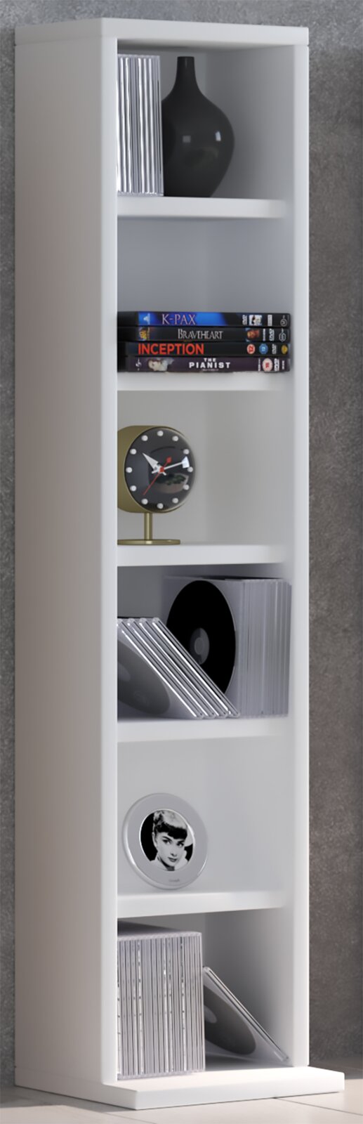 Schmales weißes Standregal mit mehreren Fächern, bestückt mit CDs, DVDs, schwarzer Vase und kleiner Tischuhr, in leichter Seitenansicht von vorn vor grauer Wand.