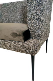 Gepolsterter Sessel mit grauem Bouclé-Bezug, schwarzem Metallfuß und beigem Kissen, Detailansicht aus schräger Seitenperspektive