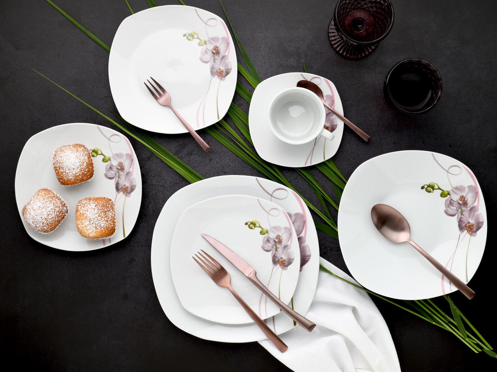 Draufsicht auf ein weißes, quadratisch abgerundetes Porzellan-Geschirrset mit Orchideen-Dekor (Teller, Schale und Tasse) samt roségoldenem Besteck, arrangiert auf dunkler Tischfläche mit grünen Blättern und Dessert.
