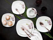 Draufsicht auf ein weißes, quadratisch abgerundetes Porzellan-Geschirrset mit Orchideen-Dekor (Teller, Schale und Tasse) samt roségoldenem Besteck, arrangiert auf dunkler Tischfläche mit grünen Blättern und Dessert.