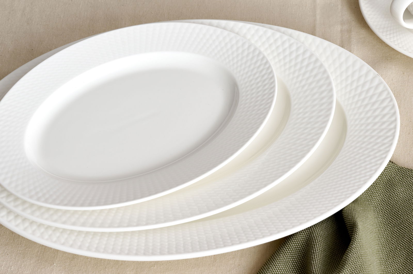 Draufsicht auf gestapelte weiße Dessertteller mit feiner Struktur auf einem Tisch mit beigem Tischtuch und grünem Stoff.