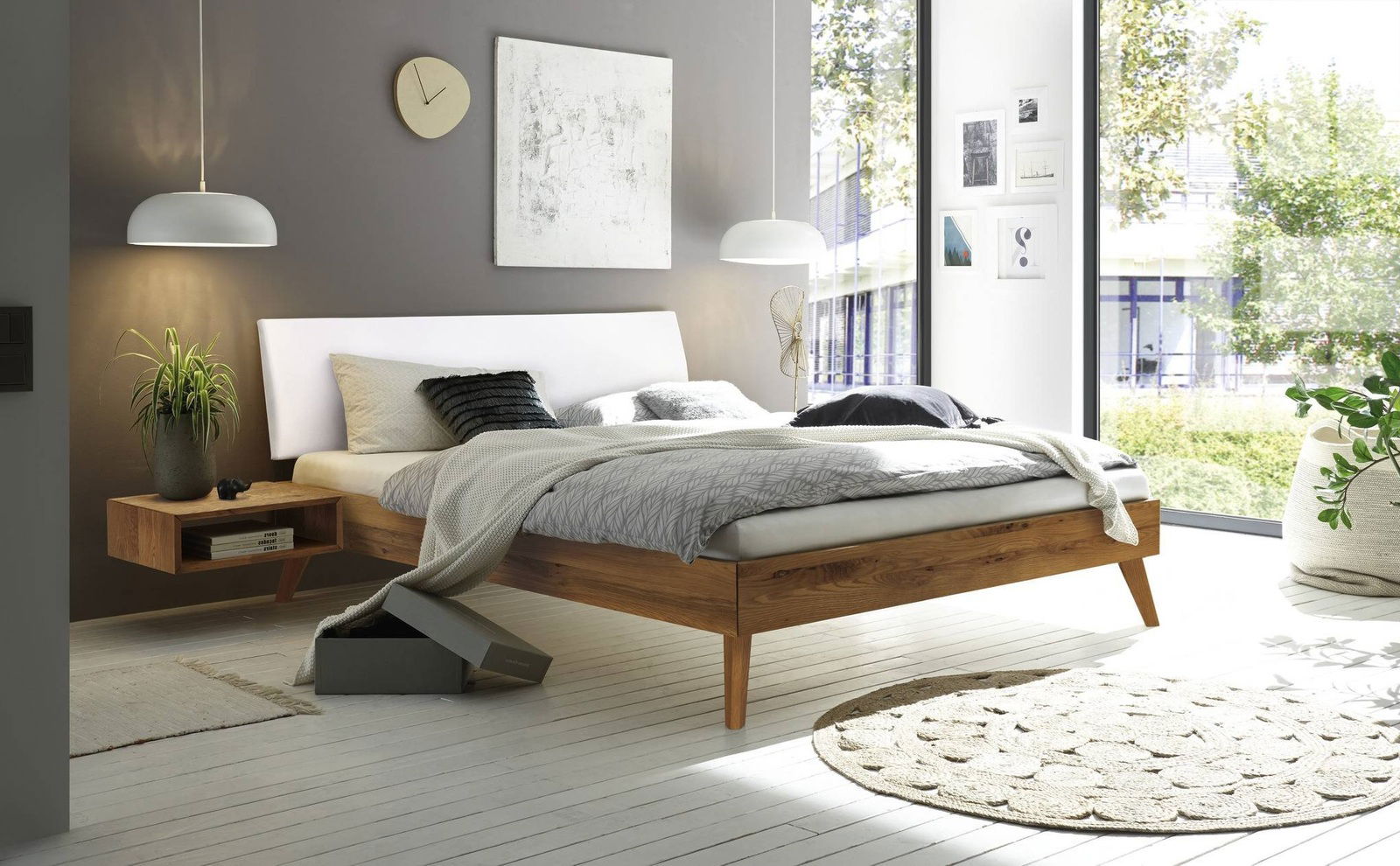 Modernes Bettgestell aus Holz mit weißem Kopfteil, seitliche Perspektive, in einem hellen Schlafzimmer mit großen Fenstern und stilvoller Dekoration.