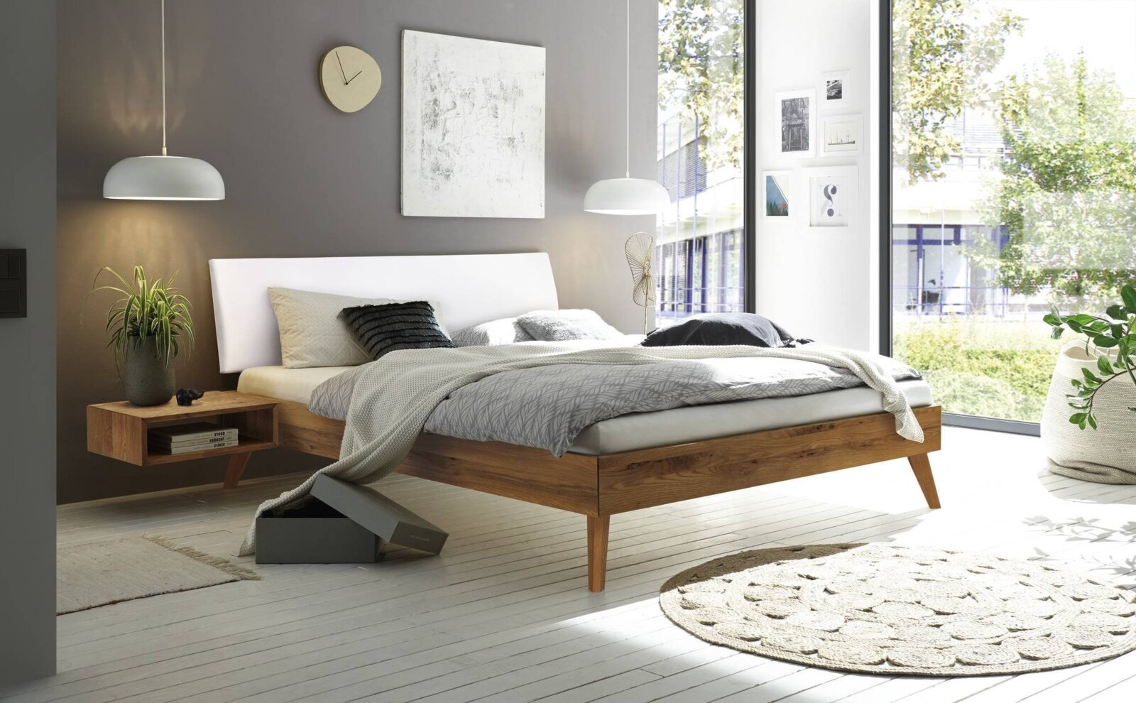 Modern houten bedframe met wit hoofdeinde, gezien vanaf de zijkant, in een lichte slaapkamer met grote ramen en een stijlvolle inrichting.