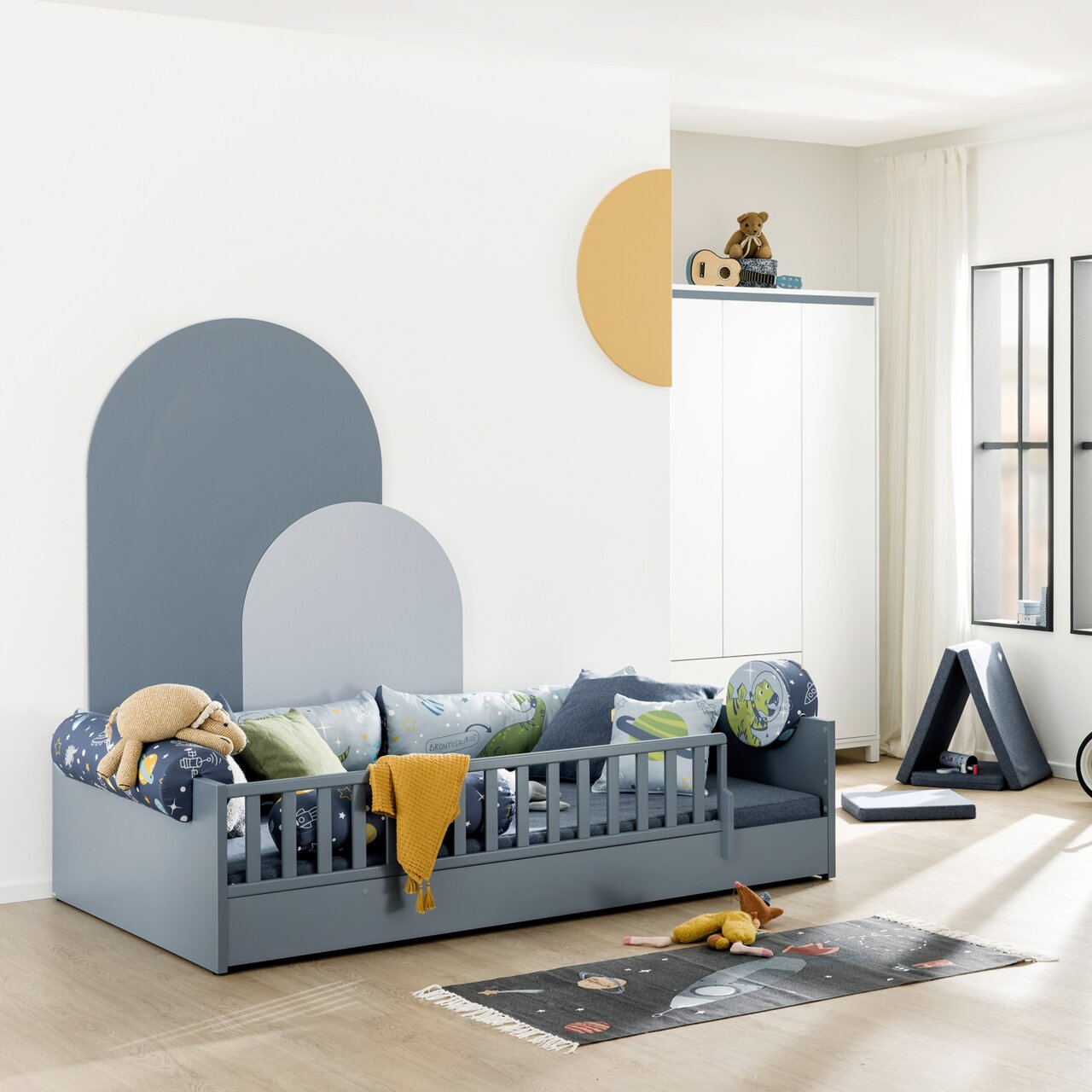 Paidi Bodenbett LITTLE FLO Kinderzimmer mit einem grauen Bodenbett mit Rausfallschutz, dekoriert mit bunten Kissen und einem gelben Tuch, seitliche Perspektive.