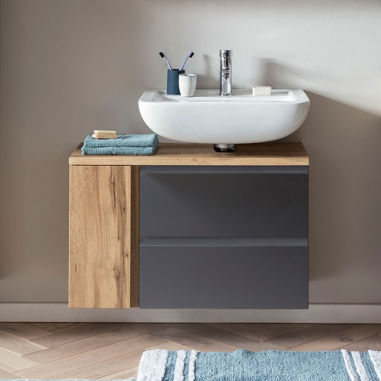 Moderner Waschbeckenunterschrank in Holzoptik mit grauen Schubladen, darauf ein weißes Waschbecken mit Armatur, von vorne betrachtet.