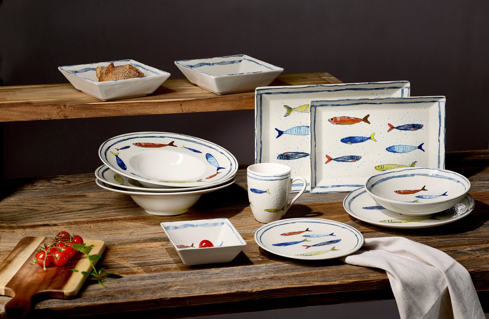 Maritimes Geschirrset aus weißer Keramik mit bunten Fischmotiven und blauen Randlinien, bestehend aus Tellern, Schalen, Tasse und eckigen Platten, in schräger Frontansicht auf einem rustikalen Holztisch arrangiert.