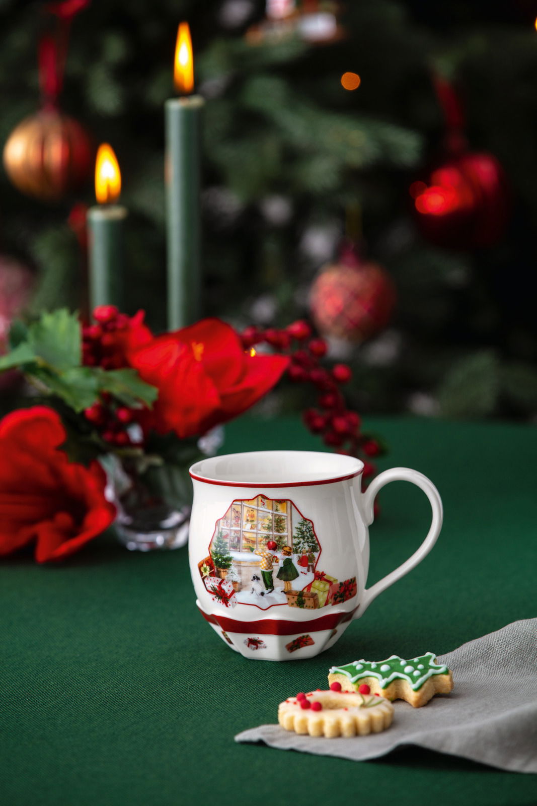 Villeroy & Boch Henkelbecher Weihnachtlich dekorierte Tasse mit Spielzeugladen-Motiv, im Vordergrund auf einem grünen Tisch mit Weihnachtsplätzchen und Kerzen im Hintergrund.