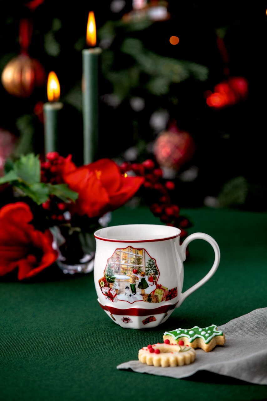 Villeroy & Boch Henkelbecher Weihnachtlich dekorierte Tasse mit Spielzeugladen-Motiv, im Vordergrund auf einem grünen Tisch mit Weihnachtsplätzchen und Kerzen im Hintergrund.
