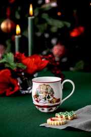 Weihnachtlich dekorierte Tasse mit Spielzeugladen-Motiv, im Vordergrund auf einem grünen Tisch mit Weihnachtsplätzchen und Kerzen im Hintergrund.
